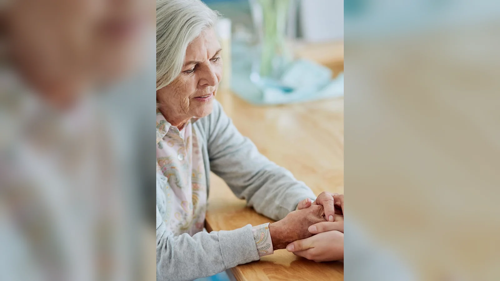 Older woman seated at a table while another person gently holds her hands in a supportive gesture